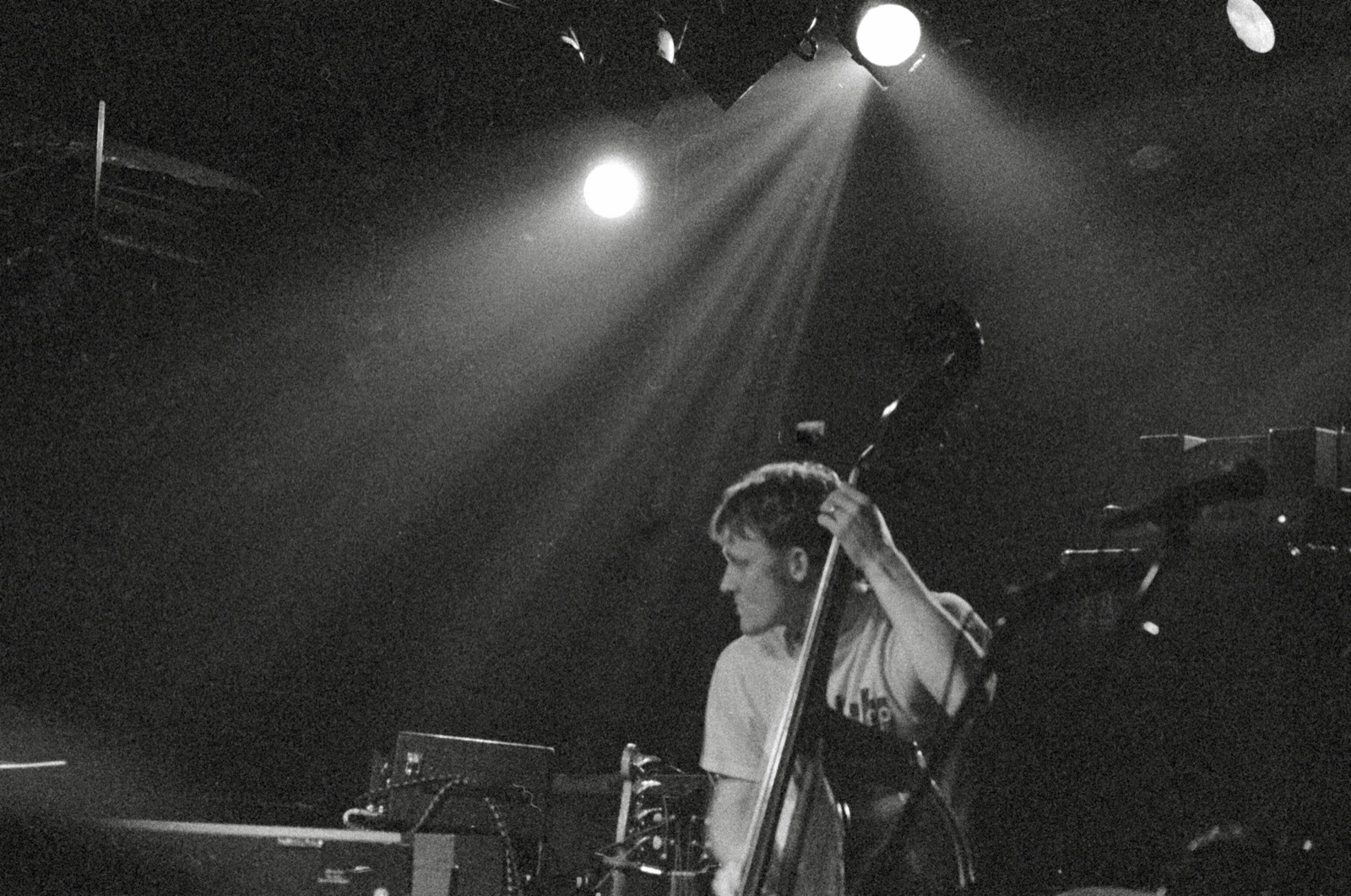 A bassist plays upright bass under spotlight beams cutting through a dark, smoky stage in black and white.